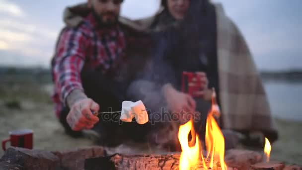 Verliefd stel zittend rond het kampvuur bij het meer, pratend, hete thee drinkend en marshmallows frituurend — Stockvideo