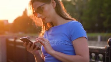 Kadın bir akıllı telefon açık havada günbatımında kullanarak güneş gözlüğü
