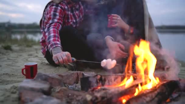 Verliebte Paare sitzen am Lagerfeuer am See, unterhalten sich, trinken heißen Tee und braten Marshmallows — Stockvideo