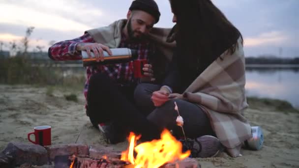 Pareja enamorada sentada alrededor de la hoguera en el lago, hablando y bebiendo té caliente — Vídeos de Stock