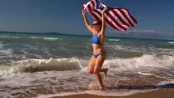 Strand bikini nő velünk zászló fut a víz mentén a tengerparton. Fogalom az Usa a függetlenség napja — Stock videók