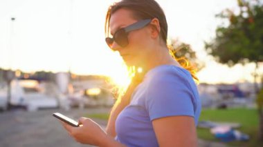Kadın bir dock yat ve tekne günbatımı, yakın çekim, bir sürü ile aşağı yürürken smartphone kullanarak güneş gözlüğü