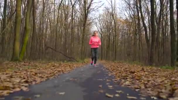 Женщина с наушниками и смартфоном бежит по осеннему лесу на закате — стоковое видео