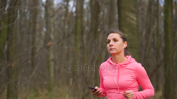 Gros plan d'une femme avec écouteurs et smartphone traversant une forêt d'automne au coucher du soleil — Video