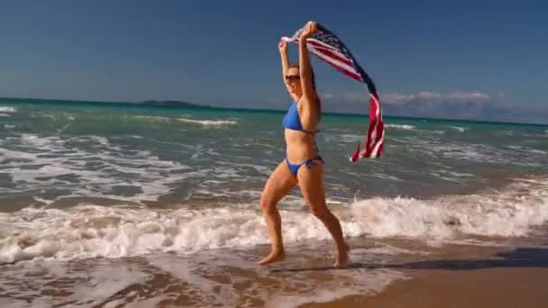Strand bikini nő velünk zászló fut a víz mentén a tengerparton. Fogalma az Usa a függetlenség napja. Lassú fogalma — Stock videók