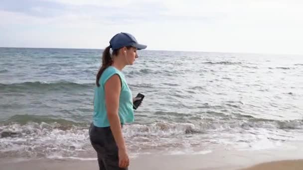 Sportieve vrouw met hoofdtelefoon wandelen langs het strand. Slow motion — Stockvideo