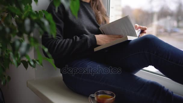 Beautiful young woman sitting on window sill with tea and reading book — Stock Video