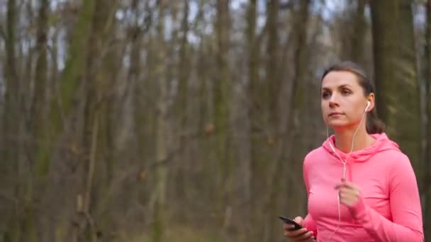 Närbild av kvinna med hörlurar och smartphone som kör genom en hösten skogen vid solnedgången. Slow motion — Stockvideo