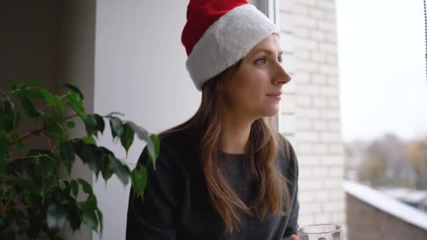 Hermosa joven en el sombrero de Santa Claus sentado en el alféizar de la ventana con té y libro de lectura en anticipación de la Navidad — Vídeos de Stock