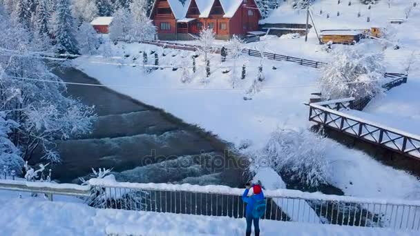 Vlucht over meisje maakt foto door smartphone op de brug over een rivier berg in de buurt van een berg bedekt met naaldhout bos in de winter. Heldere zonnige frosty — Stockvideo