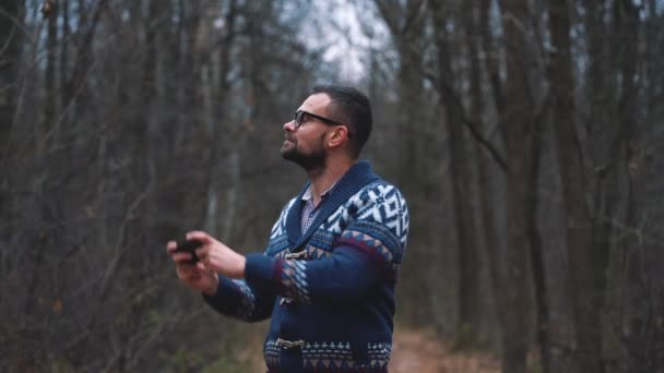 Человек в очках идет по осеннему лесу и делает фото со смартфона. Медленное движение — стоковое видео