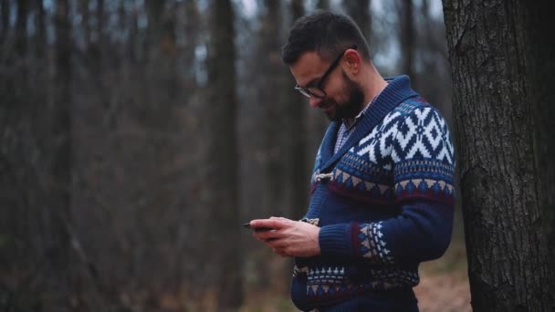 Человек в очках ходит по осеннему лесу и пользуется смартфоном. Медленное движение — стоковое видео