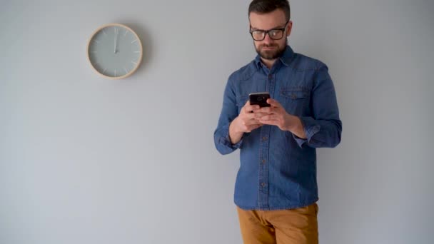 Hombre barbudo usando teléfono inteligente contra una pared gris con un reloj — Vídeos de Stock