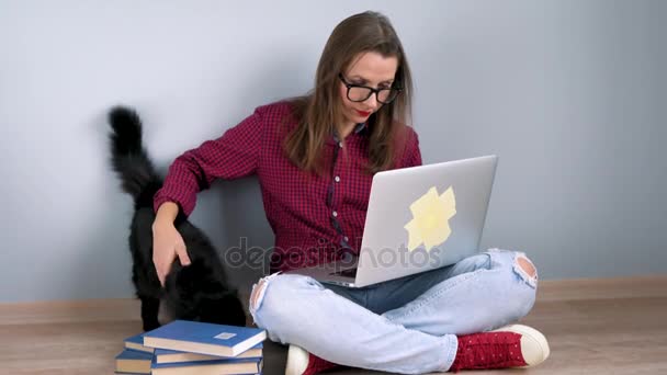 Hermosa chica usando el ordenador portátil y acariciando a un gato negro mientras está sentado en el suelo en casa — Vídeos de Stock