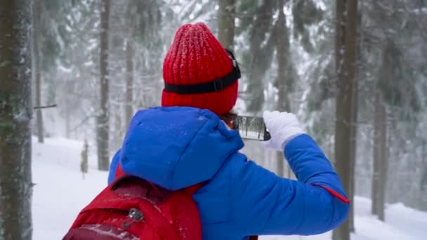 Ein einsames Touristenmädchen spaziert durch einen schneebedeckten Nadelwald in den Bergen und macht ein Foto von der Natur mit dem Smartphone. frostiges Wetter — Stockvideo