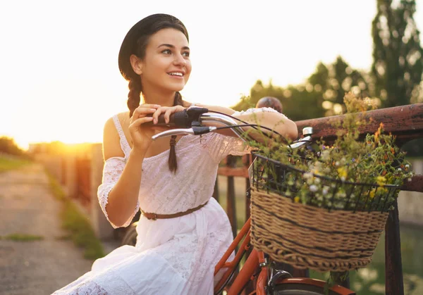 Jovem bela mulher sentada em sua bicicleta com flores ao sol — Fotografia de Stock