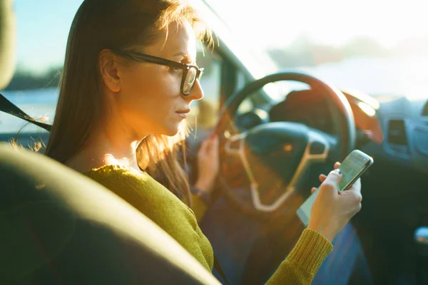 Mujer en gafas utiliza un teléfono inteligente mientras conduce un coche al atardecer — Foto de Stock