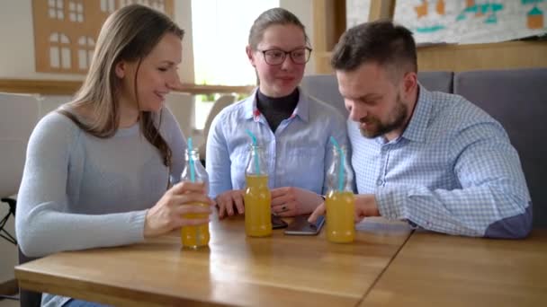 Três amigos se sentam em um café, bebem suco e se divertem se comunicando — Vídeo de Stock