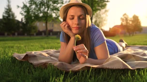 Girl with a dandelion in her hands relaxes lying down on a blanket in the park at sunset — Stock Video