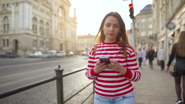Mujer joven caminando por una calle vieja usando smartphone al atardecer. Comunicación, redes sociales, concepto de compras en línea . — Vídeo de stock