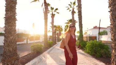 Mutlu bir kadın gün batımında palmiye sokağında hoplayıp zıplıyor ve büyük sarı bir şapka sallıyor. Yavaş çekim