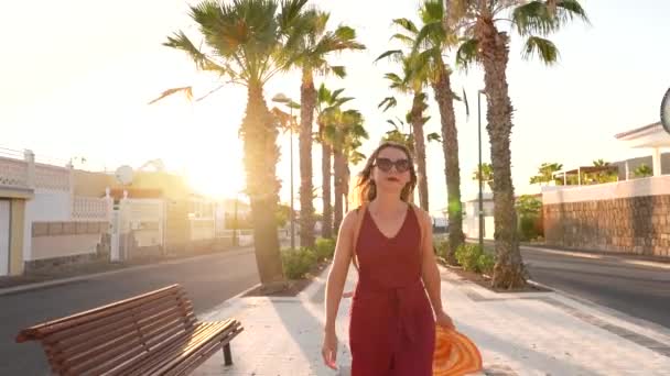 Šťastná žena ve velkém žlutém klobouku chodící po palmové uličce při západu slunce — Stock video