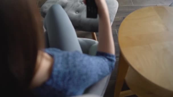 La mujer está sentada en una silla, viendo la televisión con una pantalla verde, cambiando los canales con un control remoto. Clave de croma. En interiores — Vídeos de Stock