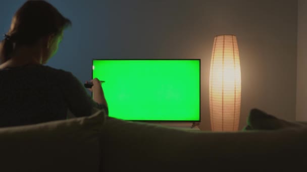 Mulher está sentada no sofá, assistindo TV com tela verde, trocando canais com controle remoto. Chave Chroma. Interior — Vídeo de Stock