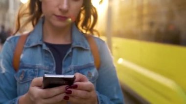 Gün batımında akıllı telefonla yaşlı bir caddede yürüyen genç bir kadın. Arka planda sarı tramvay var. İletişim, sosyal ağlar, online alışveriş kavramı.