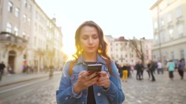 Gün batımında akıllı telefonla yaşlı bir caddede yürüyen genç bir kadın. İletişim, sosyal ağlar, online alışveriş kavramı.