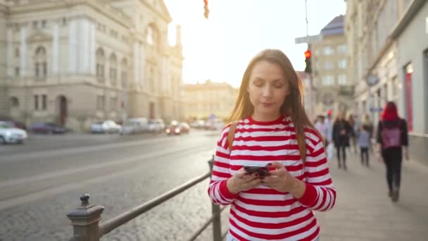 Mujer joven caminando por una calle vieja usando smartphone al atardecer. Comunicación, redes sociales, concepto de compras online. Movimiento lento — Vídeo de stock