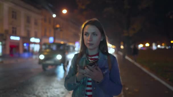 Mujer hablando en el teléfono inteligente con su amigo, lo ve y lo agita alegremente mientras camina por las calles de la ciudad nocturna — Vídeo de stock