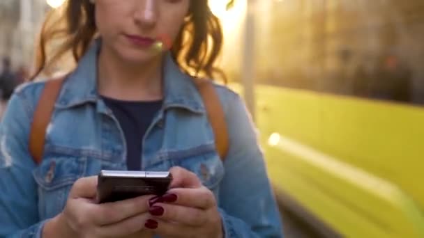 Mujer joven caminando por una calle vieja usando smartphone al atardecer. Paseos en tranvía amarillo en el fondo. Comunicación, redes sociales, concepto de compras en línea . — Vídeo de stock