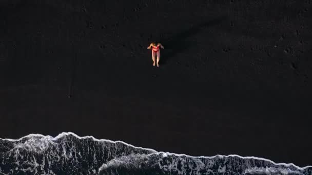 Comic video - top view of a woman in a red swimsuit who crawls along a black volcanic beach to the water to take a beautiful pose for a photo. Coast of the island of Tenerife, Canary Islands, Spain. — Stock Video