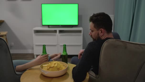 Hombre y mujer están sentados en sillas, viendo la televisión con una pantalla verde, beber cerveza, comer patatas fritas y discutir lo que ven. Vista trasera. Clave de croma. Cronograma — Vídeo de stock