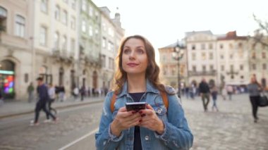 Gün batımında akıllı telefon kullanarak eski bir caddede yürüyen bir kadın. Arka planda sarı tramvay var. İletişim, sosyal ağlar, online alışveriş konsepti. Yavaş çekim