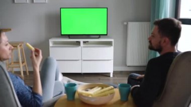 Kadın ve erkek sandalyede oturuyor, yeşil ekranlı televizyon izliyor, çay içiyor, abur cubur yiyor ve sohbet ediyorlar. Arkadan bak. Krom anahtar. Zaman Uygulaması