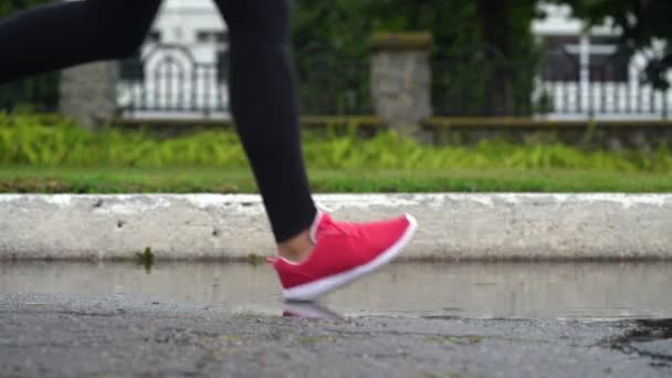 Benen van een loper in sneakers. Sportvrouw joggen buiten, stappen in modderige plas. Een hardloper die in de regen rent en plonst. Zijaanzicht. Langzame beweging — Stockvideo