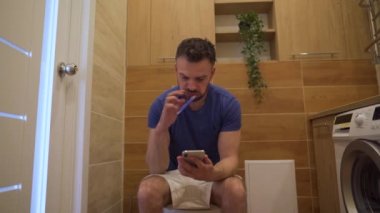 Adam tuvalette oturuyor, internette sörf yapmak için akıllı telefon kullanıyor ve aynı zamanda dişlerini tuvalette fırçalıyor.
