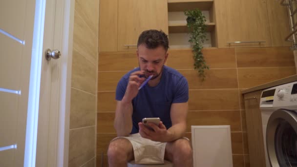 Man sitting on the toilet, uses a smartphone to surfing in Internet and brushes his teeth at the same time in restroom — Stock Video