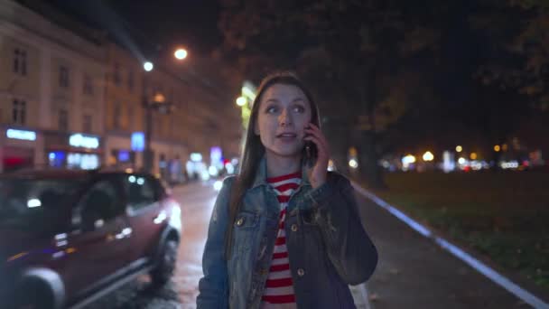 Woman talking on the smartphone with her friend, sees him on the street and calls to approach her. Blurry city lights on background — Stock Video
