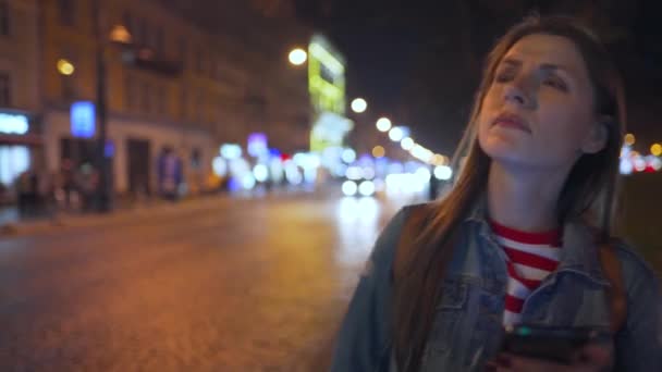 Mujer usando smartphone y considera los alrededores mientras camina por las calles de la ciudad nocturna — Vídeo de stock