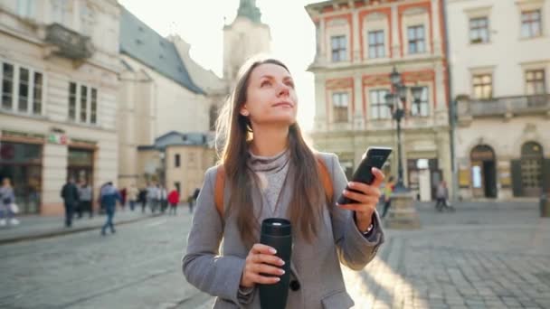 Elinde termos bardağı olan bir kadın gün batımında akıllı telefon kullanarak eski bir caddede yürüyor. İletişim, sosyal ağlar, online alışveriş kavramı. — Stok video