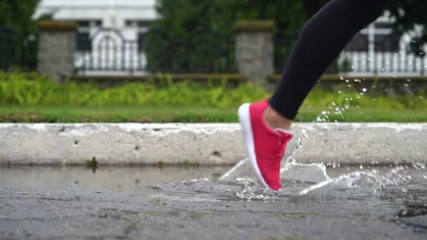 Gambe di un corridore in scarpe da ginnastica. Donna sportiva che fa jogging all'aperto, che entra in una pozzanghera fangosa. Singolo corridore che corre sotto la pioggia, facendo schizzi. Vista laterale — Video Stock