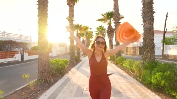 Gün batımında bir palmiye sokak boyunca yürüyen büyük sarı şapkalı mutlu kadın — Stok video