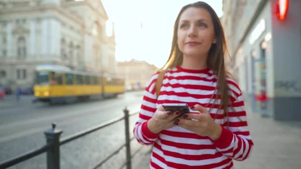 Egy nő sétál egy régi utcán okostelefonnal naplementekor. Kommunikáció, közösségi hálózatok, online vásárlási koncepció. Lassú mozgás. — Stock videók