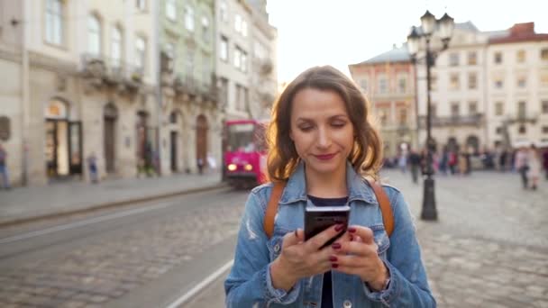 Egy nő sétál egy régi utcán okostelefonnal naplementekor. Villamos lovagol a háttérben. Kommunikáció, közösségi hálózatok, online vásárlási koncepció. Lassú mozgás. — Stock videók