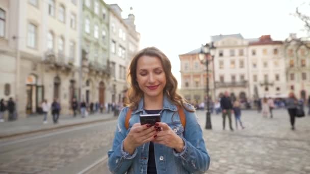 日没時にスマートフォンを使って古い通りを歩いている女性。黄色のトラムが背景に乗っています。コミュニケーション、ソーシャルネットワーク、オンラインショッピングの概念 — ストック動画