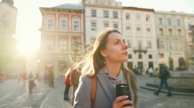 Elinde termos bardağıyla sokakta yürüyen ve soğuk bir sonbahar gününde gün batımında şehrin mimarisine hayran olan bir kadın.