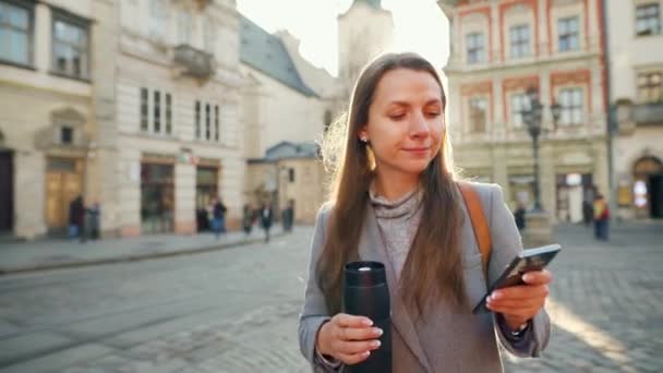 夕暮れ時にスマートフォンを使って古い通りを歩いて手に魔法瓶カップを持つ女性。コミュニケーション、ソーシャルネットワーク、オンラインショッピングの概念. — ストック動画